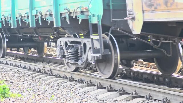 Close-up of a train wheel on railway tracks. The metal wheel is mounted on a sturdy axle, surrounded by gravel and wooden ties. The scene captures industrial transportation.
