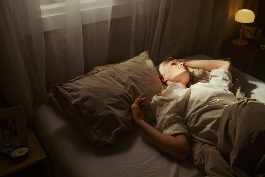 High angle view of young adult Caucasian woman lying awake in bed, touching forehead and staring upward, suggesting insomnia, stress, burnout. Useful for sleep disorder, mental health campaigns