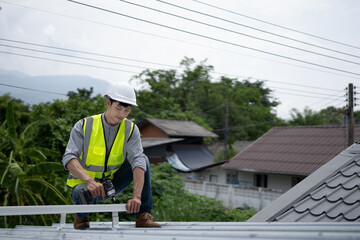 Fototapeta premium A focused Asian male technician in safety vest and hardhat uses a power drill to assemble aluminum roofing framework on a residential construction site