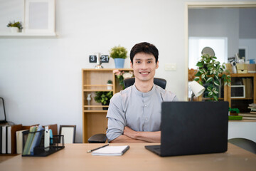 Fototapeta premium Young Asian Businessman Sitting at Desk with arms crossed and Laptop in Modern Home Office with Confident Expression, remote work and business concepts