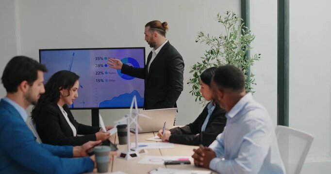 Middle Eastern businessman presents renewable energy on screen during office strategy meeting. Presenter points at turbine graphic and explains performance
