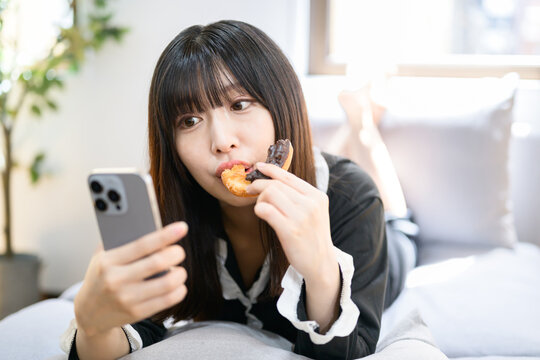 ベッドでスマホを見ながらドーナツを食べる日本人女性