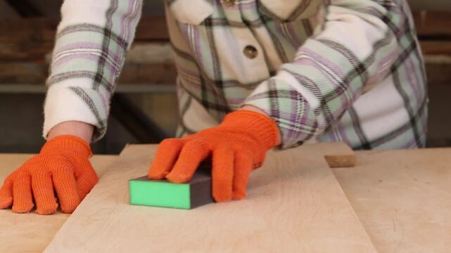 The process of sanding a wooden surface with a sanding block. The hand of a craftsman in an orange protective glove sands a wooden board with an abrasive sponge. Carpentry and wood preparation