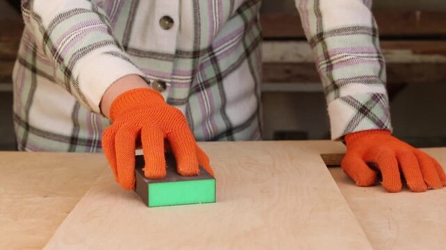 The process of sanding a wooden surface with a sanding block. The hand of a craftsman in an orange protective glove sands a wooden board with an abrasive sponge. Carpentry and wood preparation
