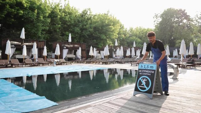 Employee carrying a safety sign with depth and no jumping rule by the swimming pool, ensuring guest safety at resort