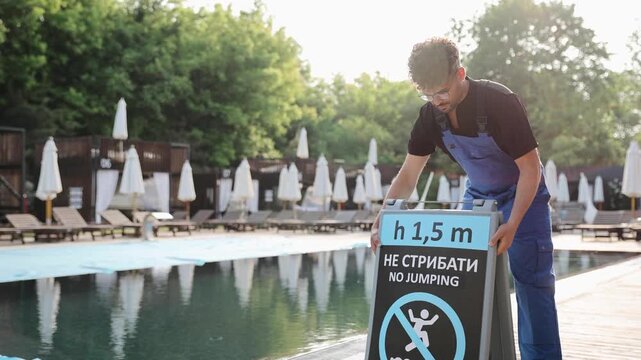Man carrying a no jumping warning sign near an outdoor swimming pool, prioritizing safety regulations and leisure experience for guests