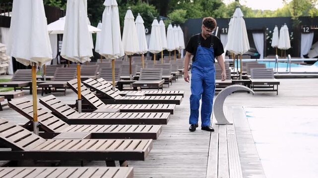 Worker in blue overalls walking on a wooden deck surrounded by sunlounge chairs and closed umbrellas, ready for morning tasks