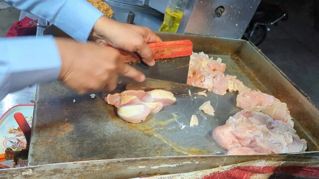 Street vendor using sharp knives to chop raw chicken meat into small pieces on a hot metal griddle for cooking delicious traditional meals in slow motion