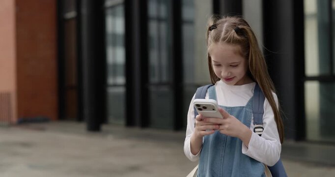 Portrait of cute little girl with schoolbag watches video via smartphone as stands on street. Happy junior schoolgirl sends message to mother outdoors