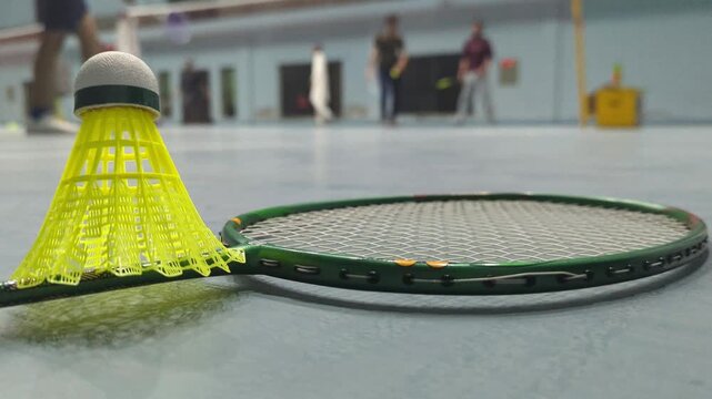 Green badminton racket lying on the indoor court floor with a bright yellow shuttlecock resting beside it while players move in the blurry background area