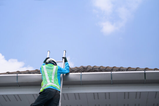 Worker in Safety Vest Climbing Ladder to Inspect Roof Gutter on Residential House Against Blue Sky