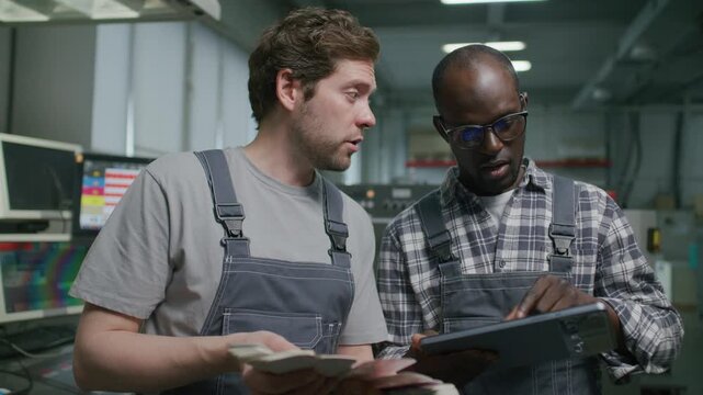 Caucasian and African American workers reviewing printed color samples, using digital tablet and talking beside industrial press in printing house