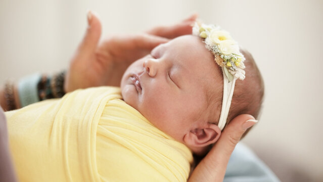Baby Wrapped In Yellow Cloth Lies Peacefully In Photographer Hands. Flower Headband Adds Charm. Warm Moment Captured Indoors. Newborn Photo Shoot.