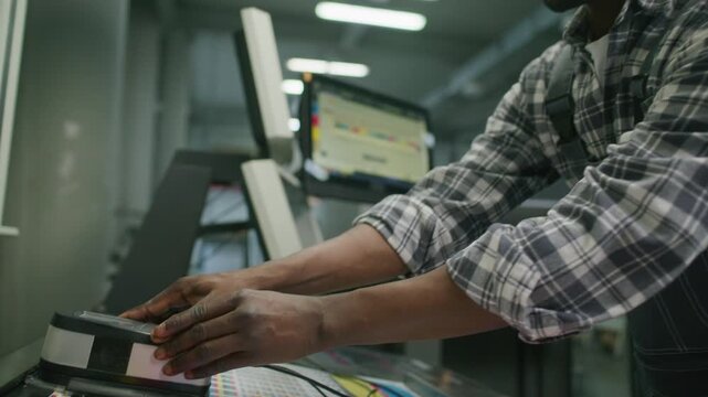 African American printing press operator scanning printed color control chart with spectrophotometer in printing house. Tilt down, close up view