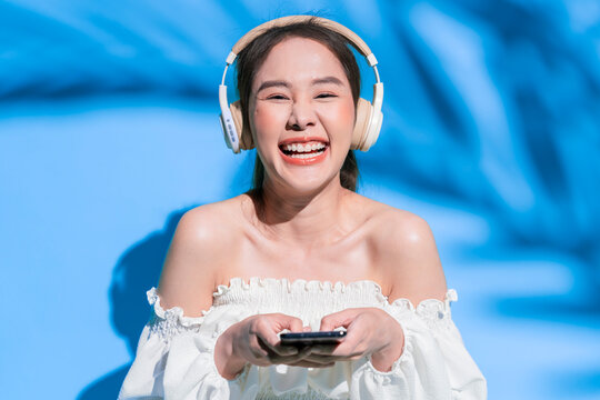 A beautiful cheerful young Asian woman wearing a trendy white off-the-shoulder crop top and wireless headphones laughs joyfully at the camera while holding a smartphone amidst tropical palm leaf shado