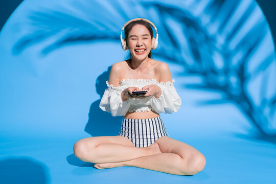 A beautiful cheerful young Asian woman wearing a trendy white off-the-shoulder crop top and wireless headphones laughs joyfully at the camera while holding a smartphone amidst tropical palm leaf shado
