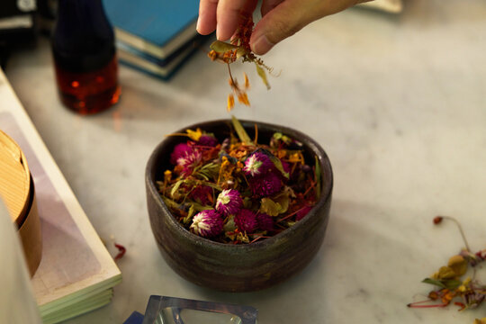 Close-up of hand sprinkling dried herbal flowers and botanicals into a ceramic bowl for tea or aromatherapy.