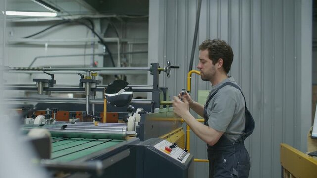 Young male printing technician in workwear operating industrial press machine in modern printing factory, manually setting up rollers and using control panel