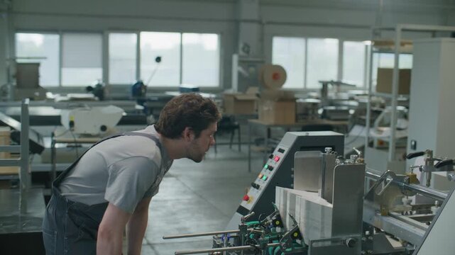 Technician in overalls pressing button on control panel, examining stack of paper sheets feeding into industrial printing machine and using computer to control equipment in printing factory