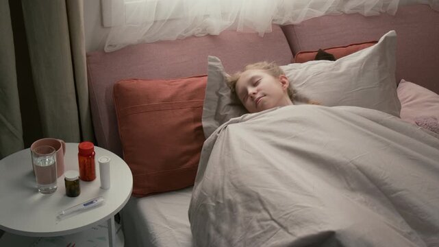 Sick teenage girl pulling up blanket while napping with plush toy in cozy bed, bottles with pills, thermometer, glass of water on white side table