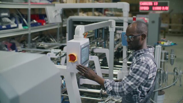 African American worker using digital touchscreen console and holding handheld electronic controller device while adjusting modern automated machinery in printing factory