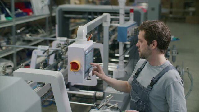 Industrial worker in overalls operating automated machinery using digital touchscreen console and handheld electronic controller in commercial printing factory