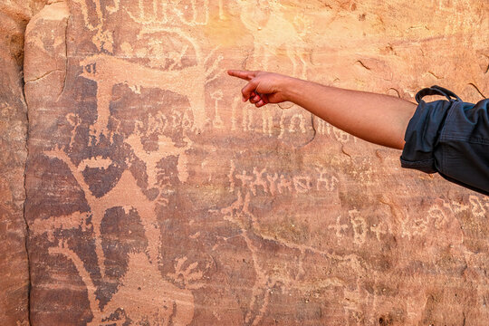 AlUla, Saudi Arabia,Jan 5, 2025: Person pointing to ancient petroglyphs of animals and human figures etched into weathered sandstone on textured rock face at Sharaan Nature Reserve, AlUla Saudi Arabia