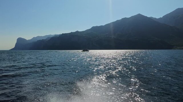 Sunlight glittering on calm lake water with distant mountain silhouettes and small boat, summer landscape scenery