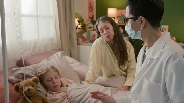 Tilt down shot of mature female physician setting intravenous infusion system for teenage patient and sitting down on bed speaking to child and her mother