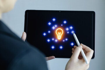 Person uses digital tablet to create light bulb design with stylus and glowing graphics in a bright workspace © yurakrasil