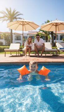 Small child wearing orange armbands swimming in pool with parents watching from lounge chair. Water safety awareness and family recreation during vacation at luxury tropical resort.