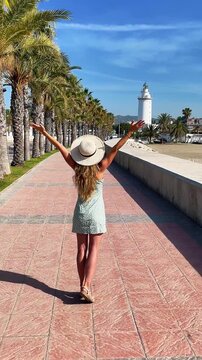 happy woman in straw hat and summer dress with arms raised standing on a promenade with palm trees looking at la farola lighthouse in malaga spain