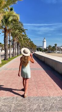 back view of a female tourist with hat and summer dress walking on promenade with palm trees toward la farola lighthouse in malaga spain