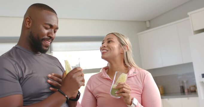 Couple in activewear laughing as man raising green smoothie glass in kitchen, toasting health