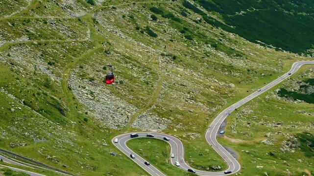 Red cable car traveling over the winding Transfagarasan mountain road in Romania. Panning camera tracks the gondola above scenic hairpin turns.