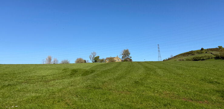 A lone house stands atop a gentle green hill beneath a clear, cloudless blue sky. The open landscape and soft light lend a quiet rural calm to the scene near Hallas Lane, Bradford, UK.