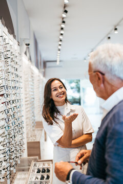 A professional female optometrist smiles while assisting a senior male customer in selecting the perfect pair of eyeglasses at a modern optical boutique.