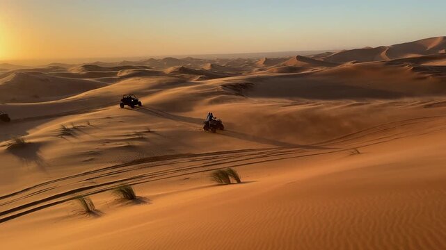 A stunning cinematic pan shot showing buggies and quad bikes driving through the endless sand dunes of Erg Chebbi under a massive golden sunset sky in Morocco