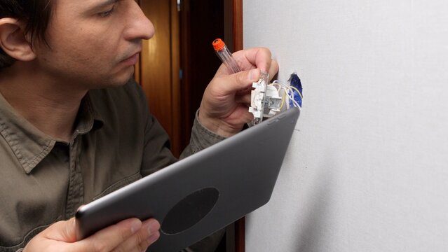 Professional electrician connecting electrical wires to a new wall socket, utilizing a digital tablet for reference or smart home integration during renovation and installation