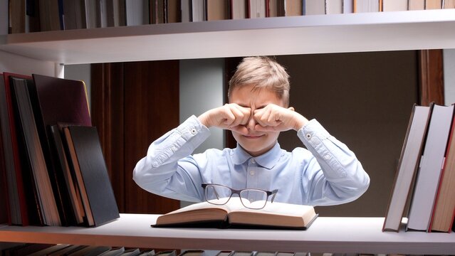 Young boy in a library rubbing dry eyes and grimacing while studying a book, showing eye strain, fatigue and discomfort from prolonged reading and schoolwork