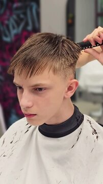 teenage boy in white barber cape with eyes closed while female barber with smartwatch trims wet hair with scissors and comb at barbershop, graffiti wall behind