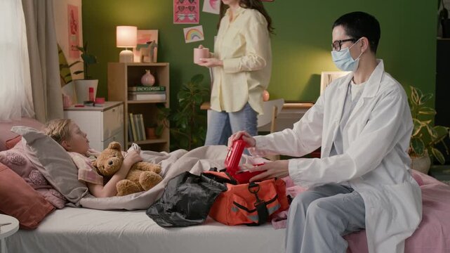 Female doctor wearing white coat and face mask looking through medicines in red first aid box while visiting sick child at home, mother bringing mug of hot tea, girl drinking it