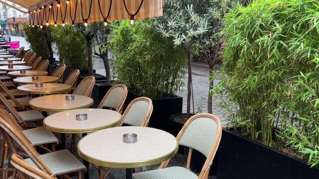 Empty terrace of Parisian cafe on spring rainy day, lush green plants surround tables and chairs on wet cobblestone streets, no people at tables covered with rainy water, cozy, atmospheric view