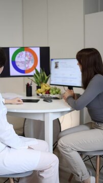 Vertical screen young woman attends modern nutrition consultation in private clinic while specialist uses digital health records and visual nutrition analytics for personalized care