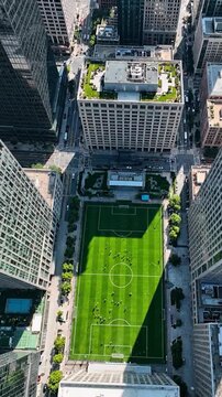 Aerial drone view looking straight down at a bright green urban football court surrounded by city skyscrapers with players moving on the field.