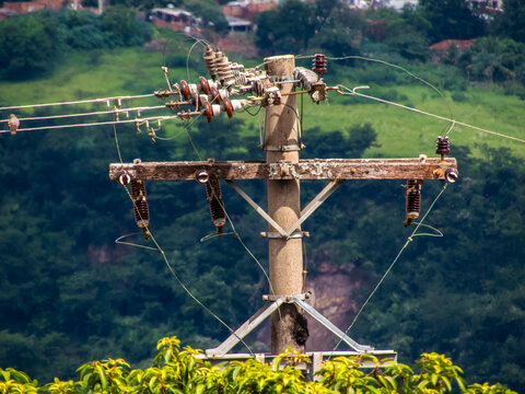 Detailed shot of an electrical distribution pole featuring weathered wooden crossarms, insulators, and power lines against a blurred natural green landscape in rural Brazil.