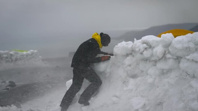 Person wearing winter gear building an igloo wall for shelter during a heavy snowstorm