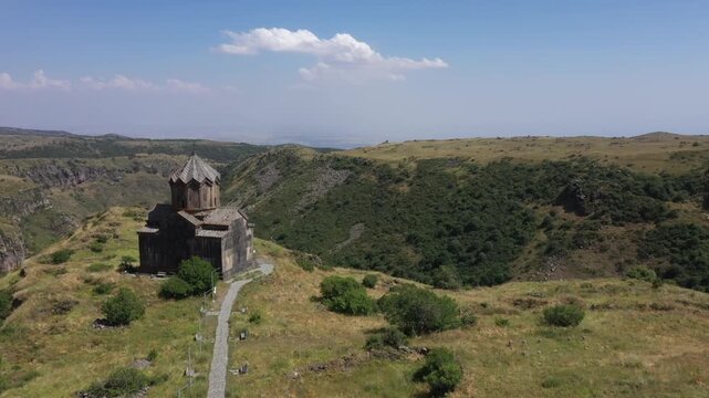 An aerial shot captures Vahramashen Church perched on a grassy hill, overlooking a dramatic gorge with mountains and a cloudy blue sky in the background.

