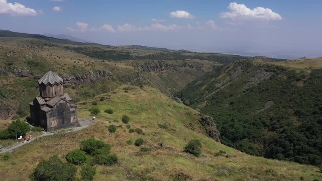 An aerial shot captures Vahramashen Church perched on a grassy hill, overlooking a dramatic gorge with mountains and a cloudy blue sky in the background.

