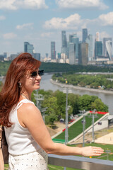 Fototapeta premium Red-haired woman overlooks Moscow skyline from observation deck near Moscow River with green parkland, modern towers under summer clouds. Tourism, city travel, lifestyle, urban panorama, social media